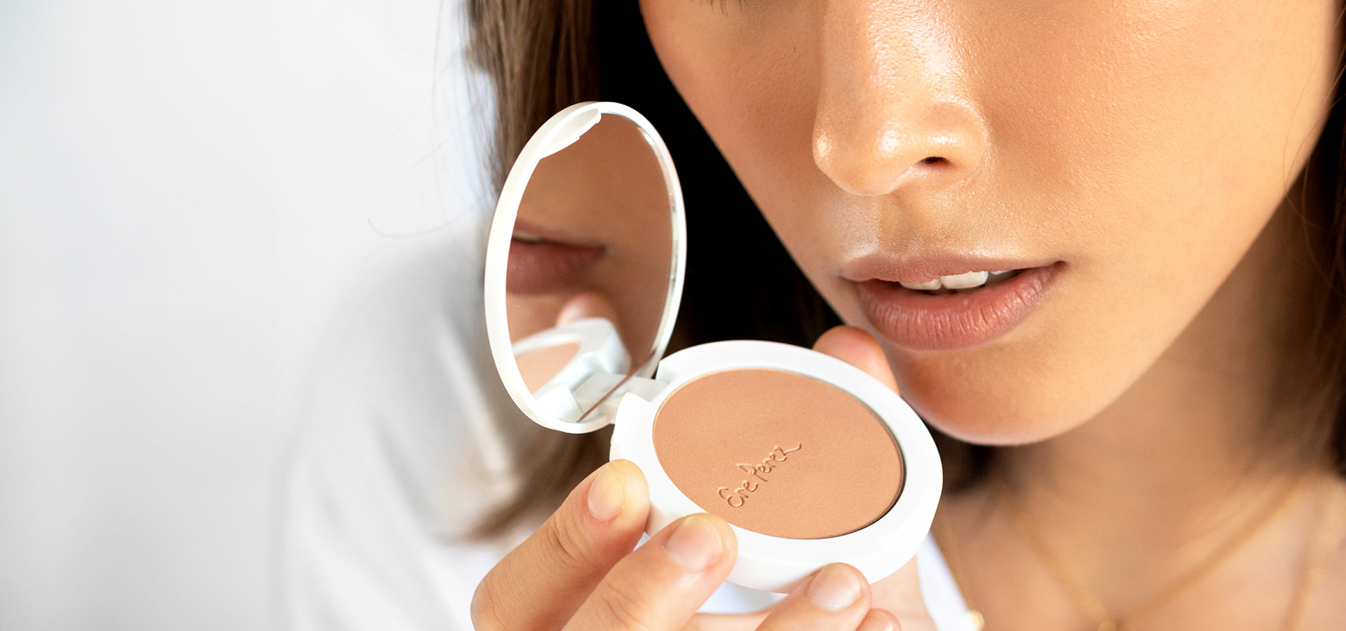 Close-up of a person holding a compact powder with a mirror, focusing on the lips and lower face. The compact has a logo embossed on the powder and features a white casing.