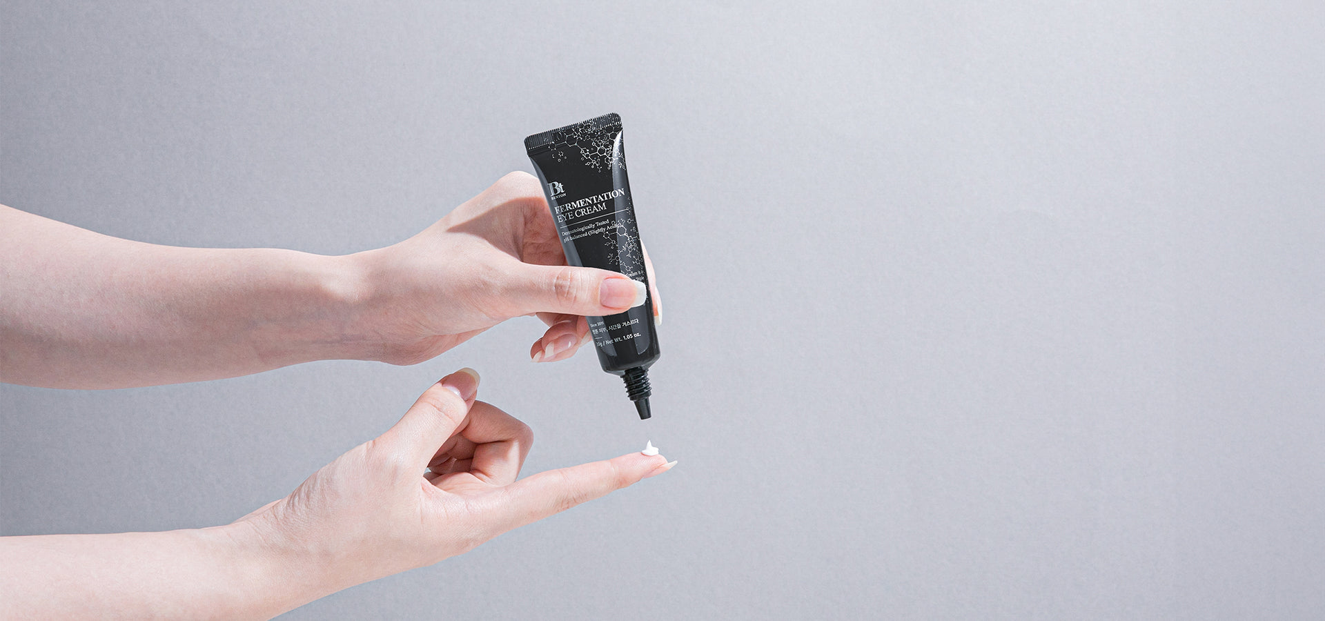 A hand squeezing a small amount of eye cream from a black tube onto a fingertip against a light gray background.