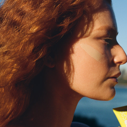 Model with sunscreen on cheek holding Mother Mousse SPF50 tube outdoors.