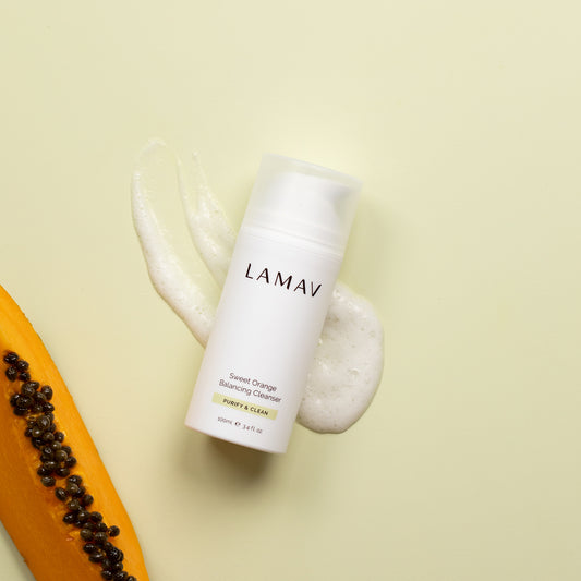 A bottle of LaMav Sweet Orange Balancing Cleanser on a light surface with foam and a slice of papaya beside it.