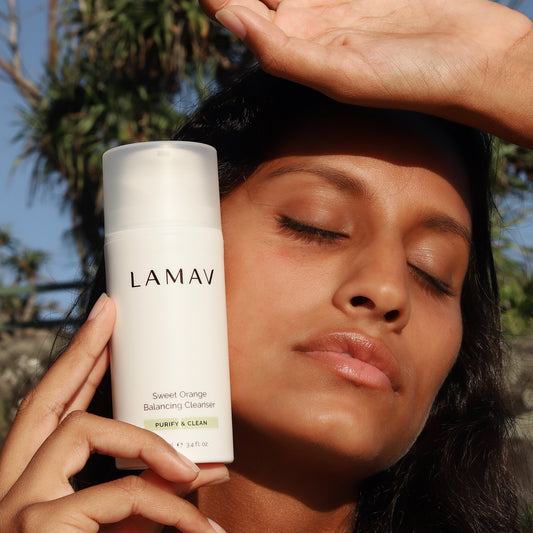 A person holding a bottle of LAMAV Sweet Orange Balancing Cleanser close to their face. The person has their eyes closed and their hand raised above their forehead. The background shows blurred greenery.