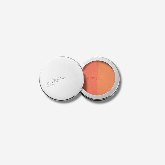 A compact of rice powder blush with an open lid, revealing two shades of blush in a sleek, silver container with a brand logo on the lid.
