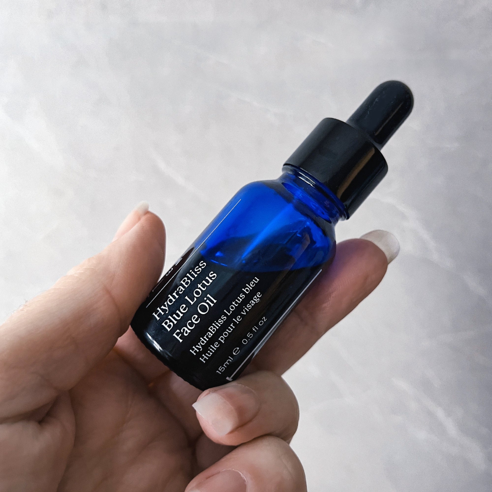 Hand holding a blue dropper bottle labeled 'HydraBliss Blue Lotus Face Oil' against a light background