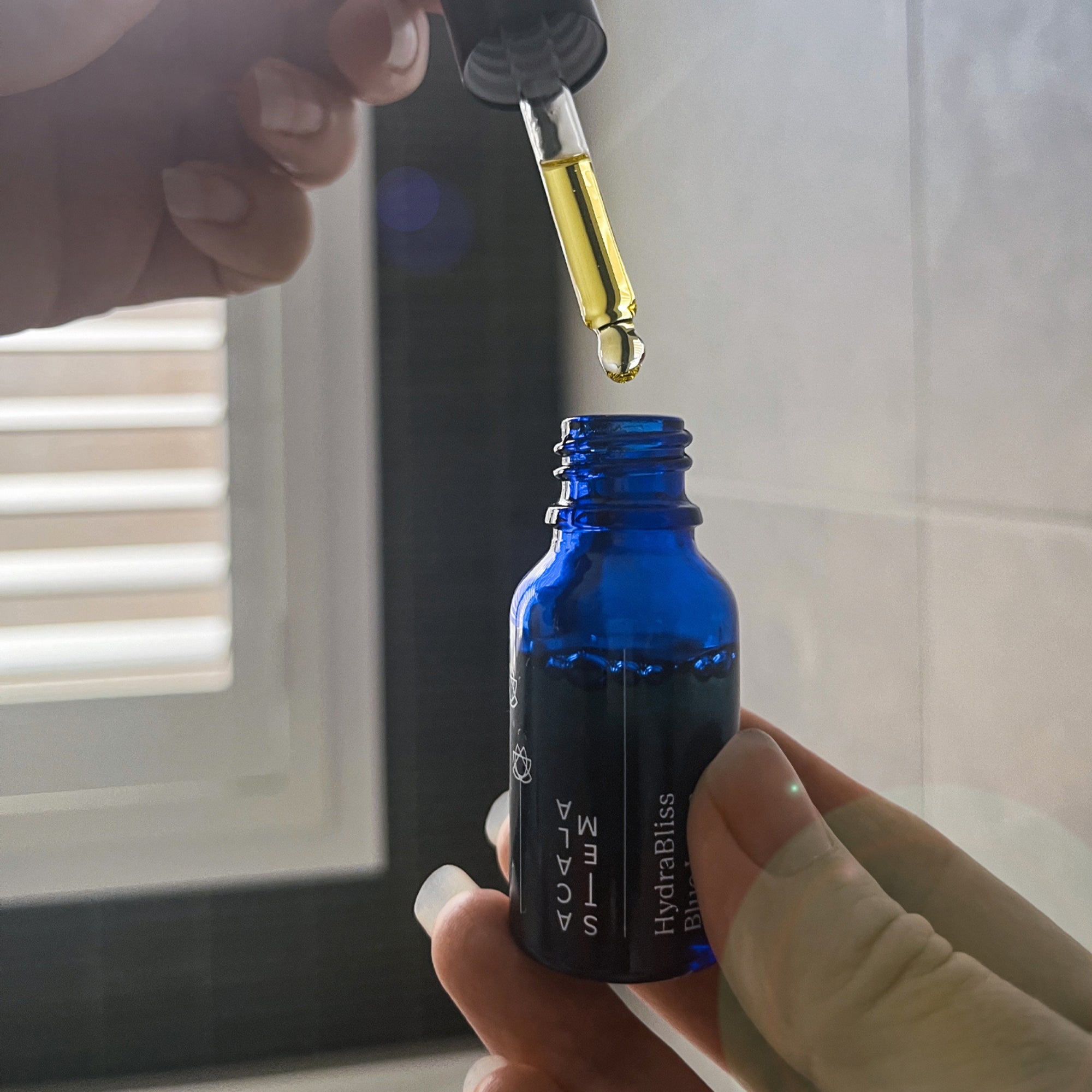 Person using a dropper on a blue bottle labeled Acala Stem Hydabliss Blue Lotus Face Oil