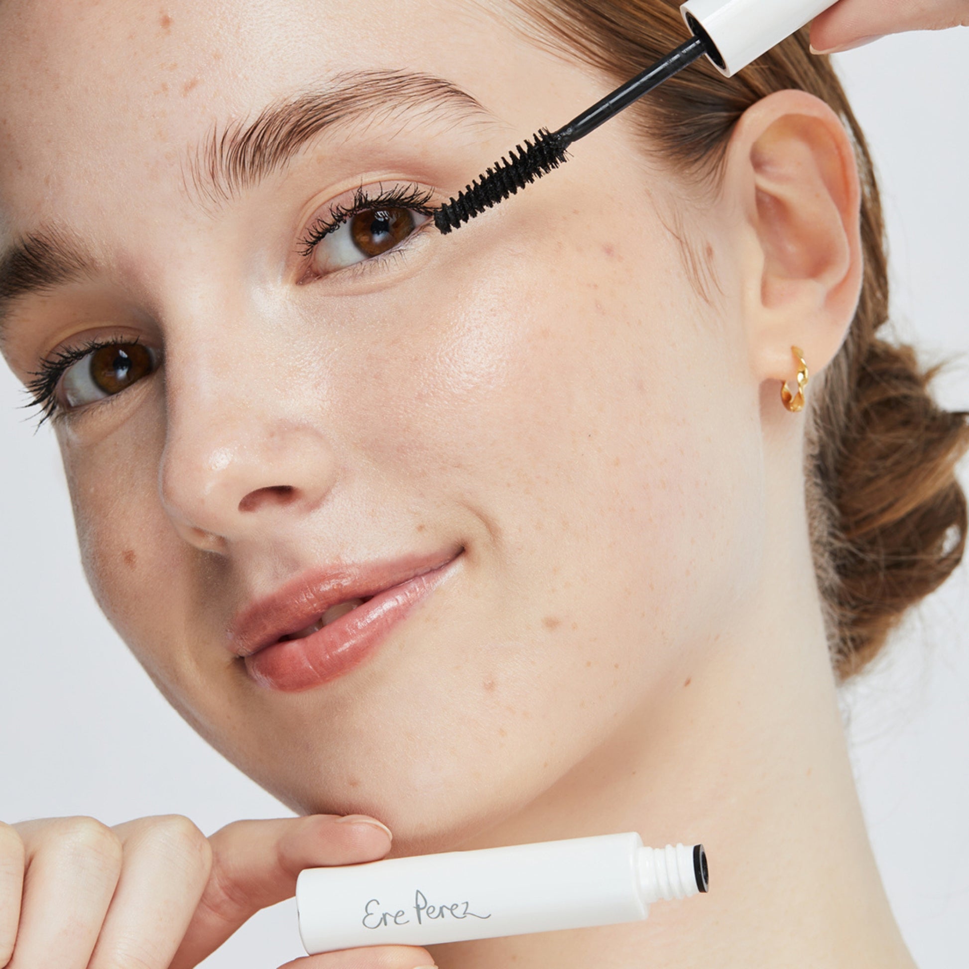 A person applying avocado waterproof mascara with an open tube labeled 'Ere Perez' visible near their face. The mascara wand is applied to the right eyelashes.