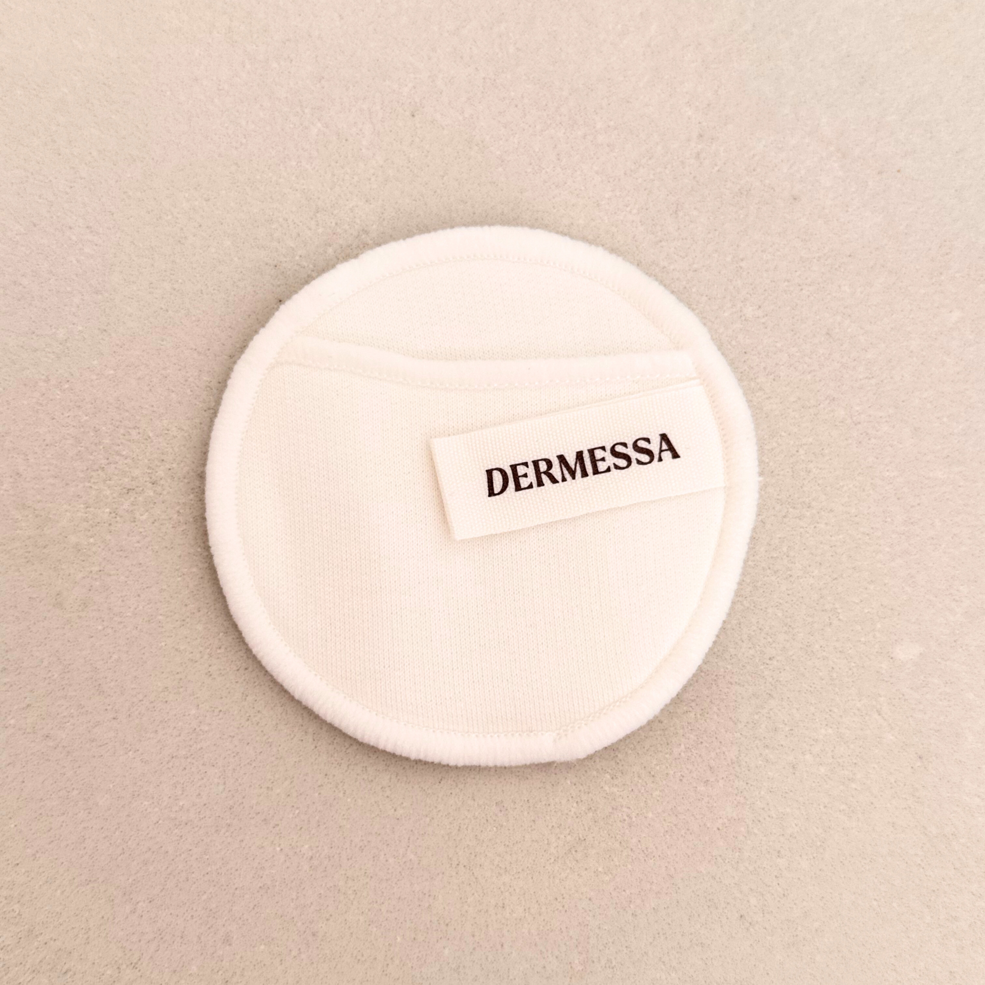 Round white makeup pad with 'DERMESSA' label on a beige background