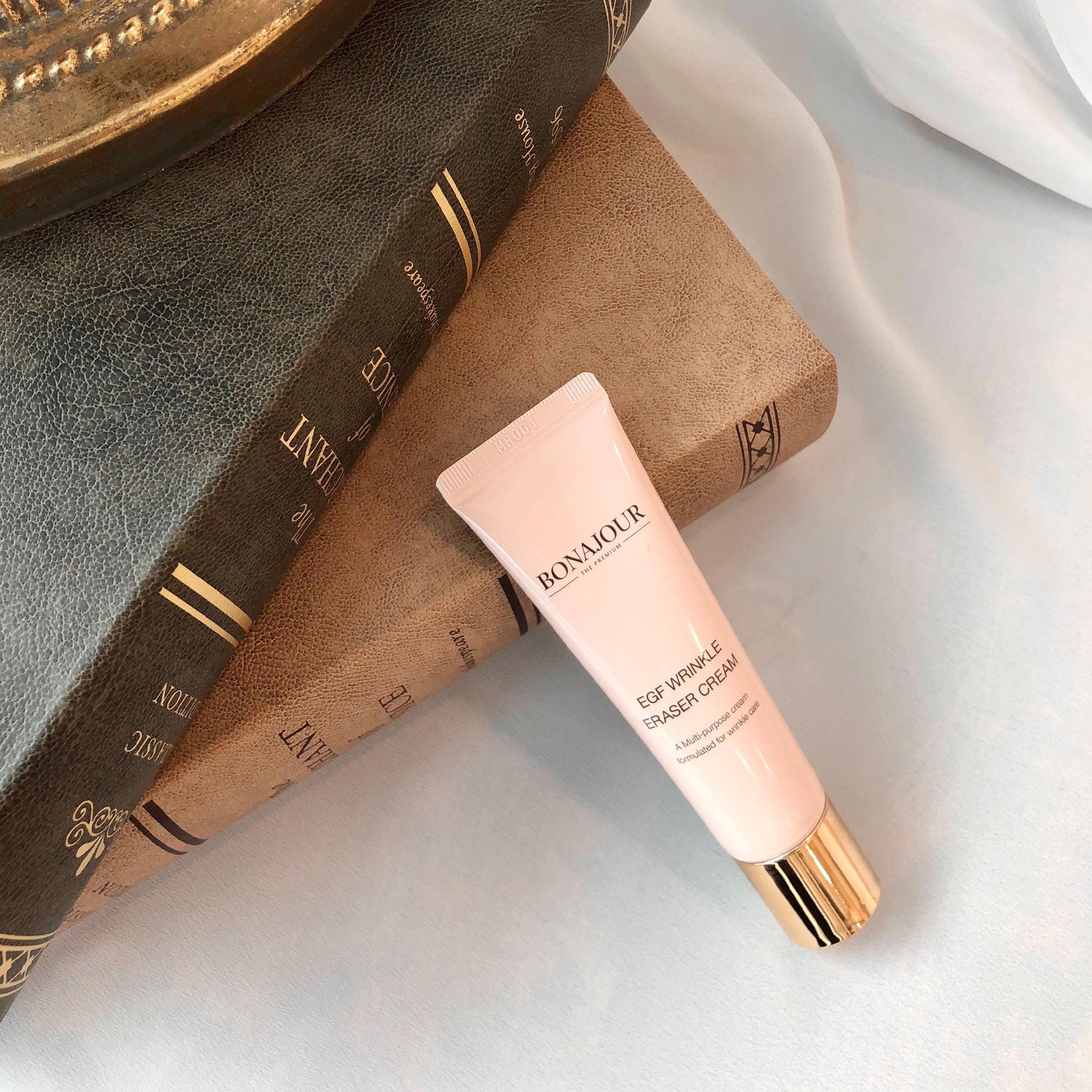 A tube of Bonajour EGF Wrinkle Eraser Cream with a gold cap placed on two stacked hardcover books with textured covers.