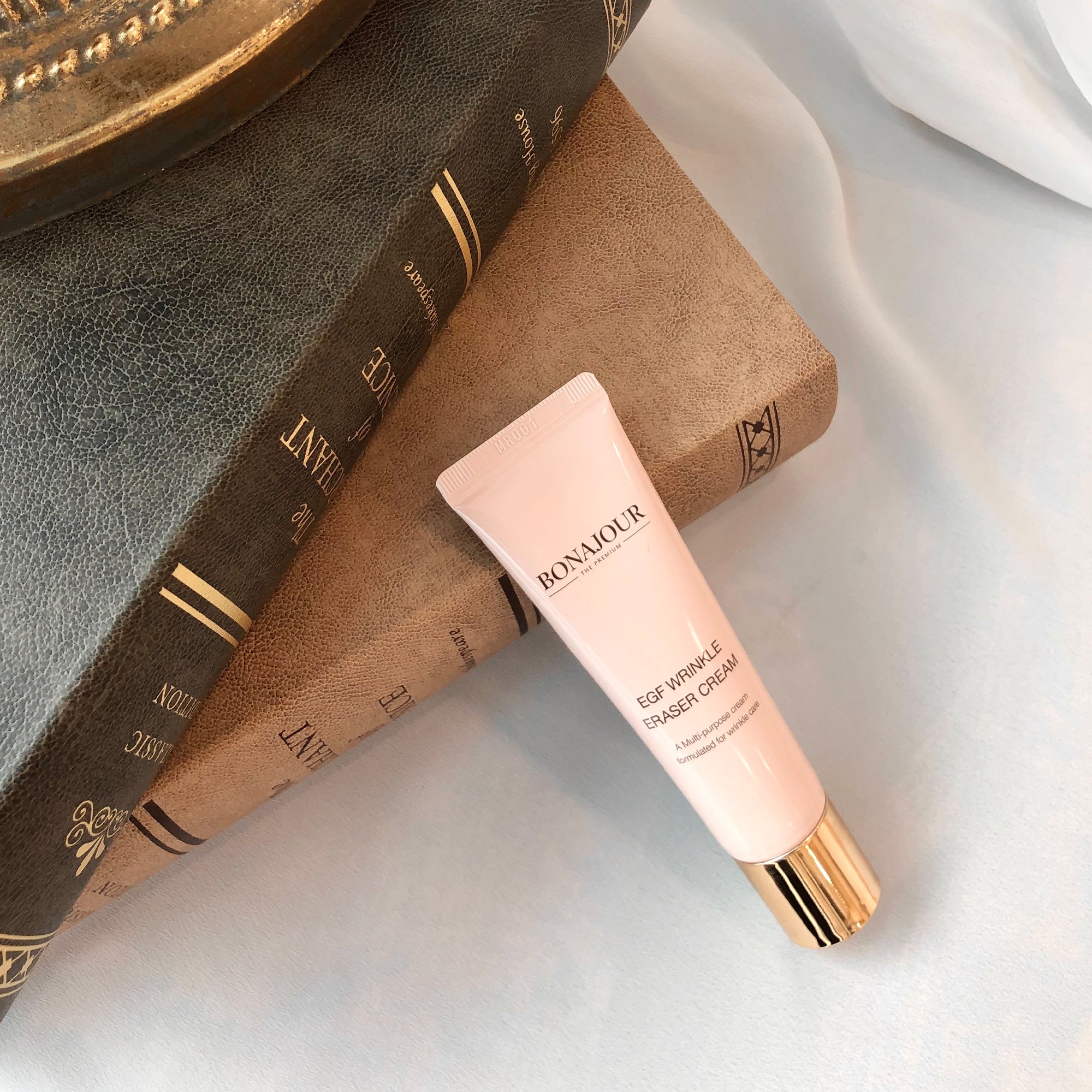 A tube of Bonajour EGF Wrinkle Eraser Cream with a gold cap placed on two stacked hardcover books with textured covers.
