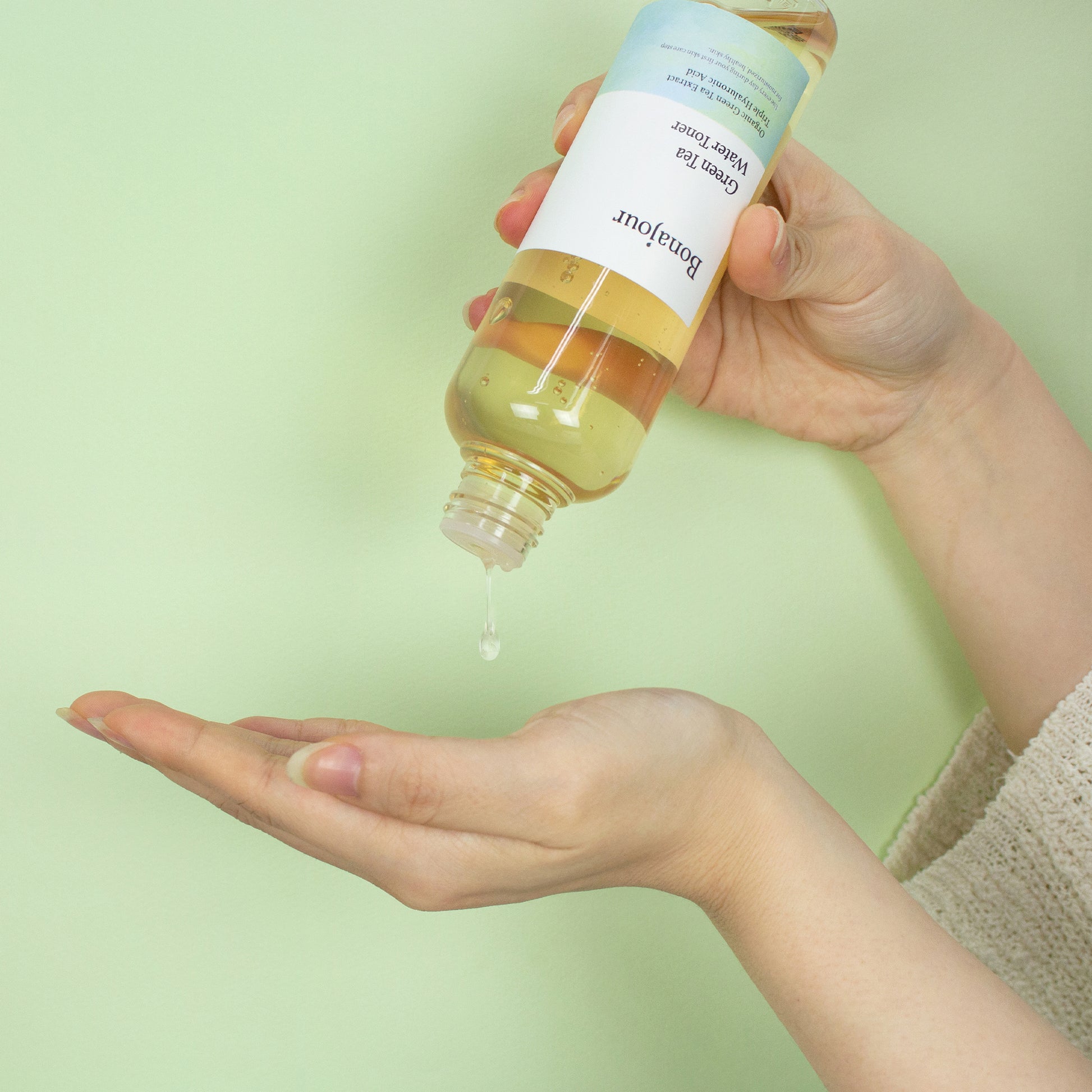 A person pouring Green Tea Water Toner from a clear bottle into their palm against a pale green background.