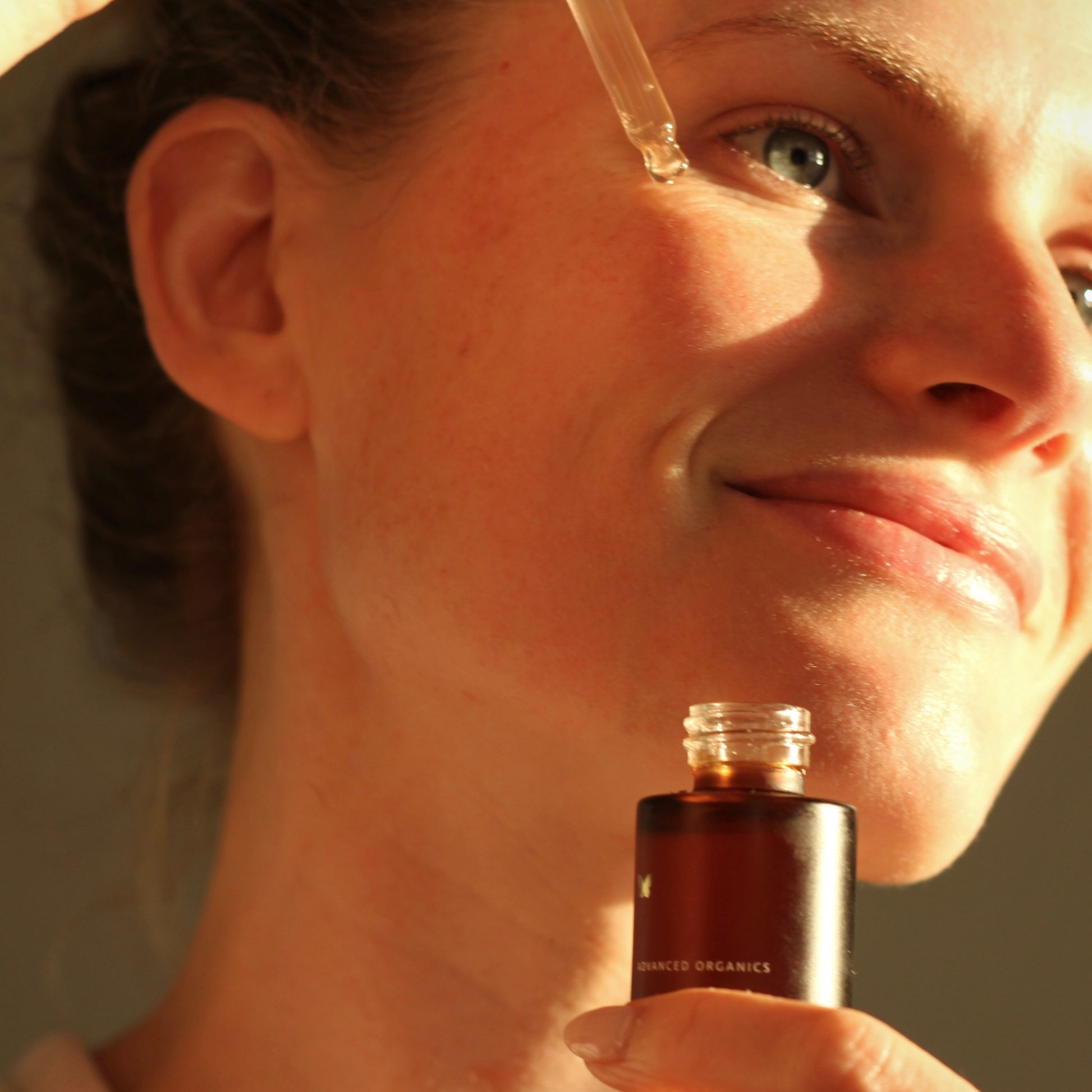 Close-up of a woman applying a dropper of liquid to her face, with a bottle of the product in front.