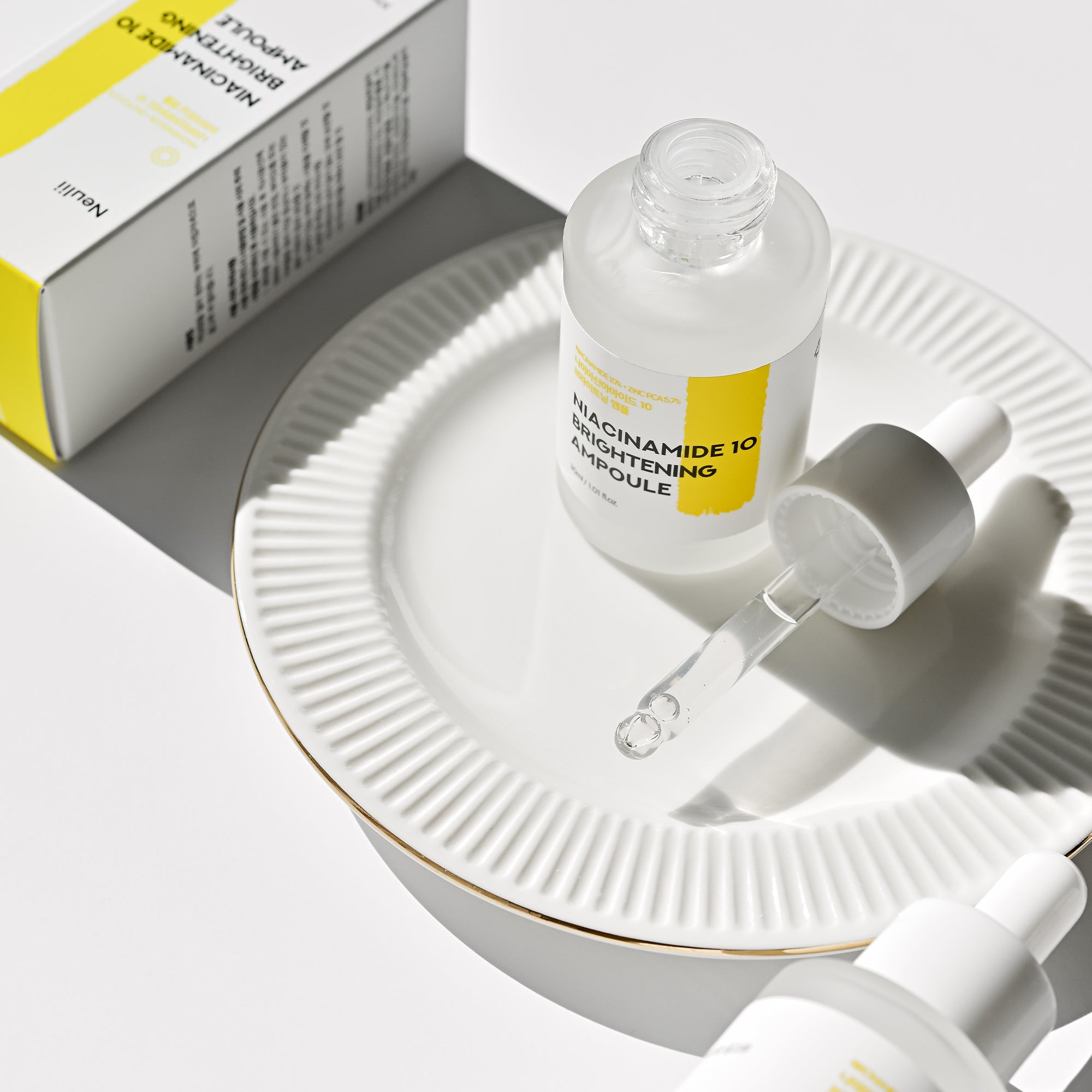 A Niacinamide 10% Brightening Ampoule bottle with a dropper, placed on a white ceramic plate, next to its packaging box.