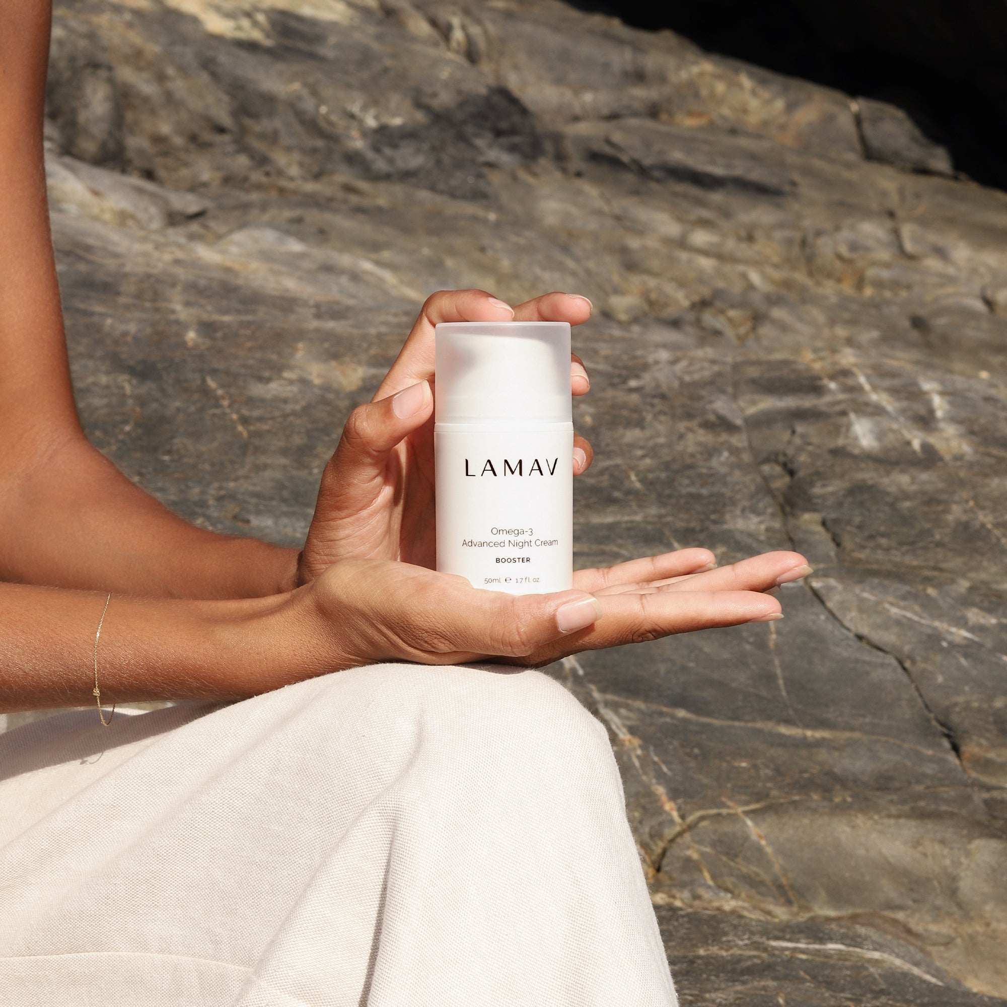 Person holding a bottle of LAMAV Omega-3 Advanced Night Cream against a rocky background.