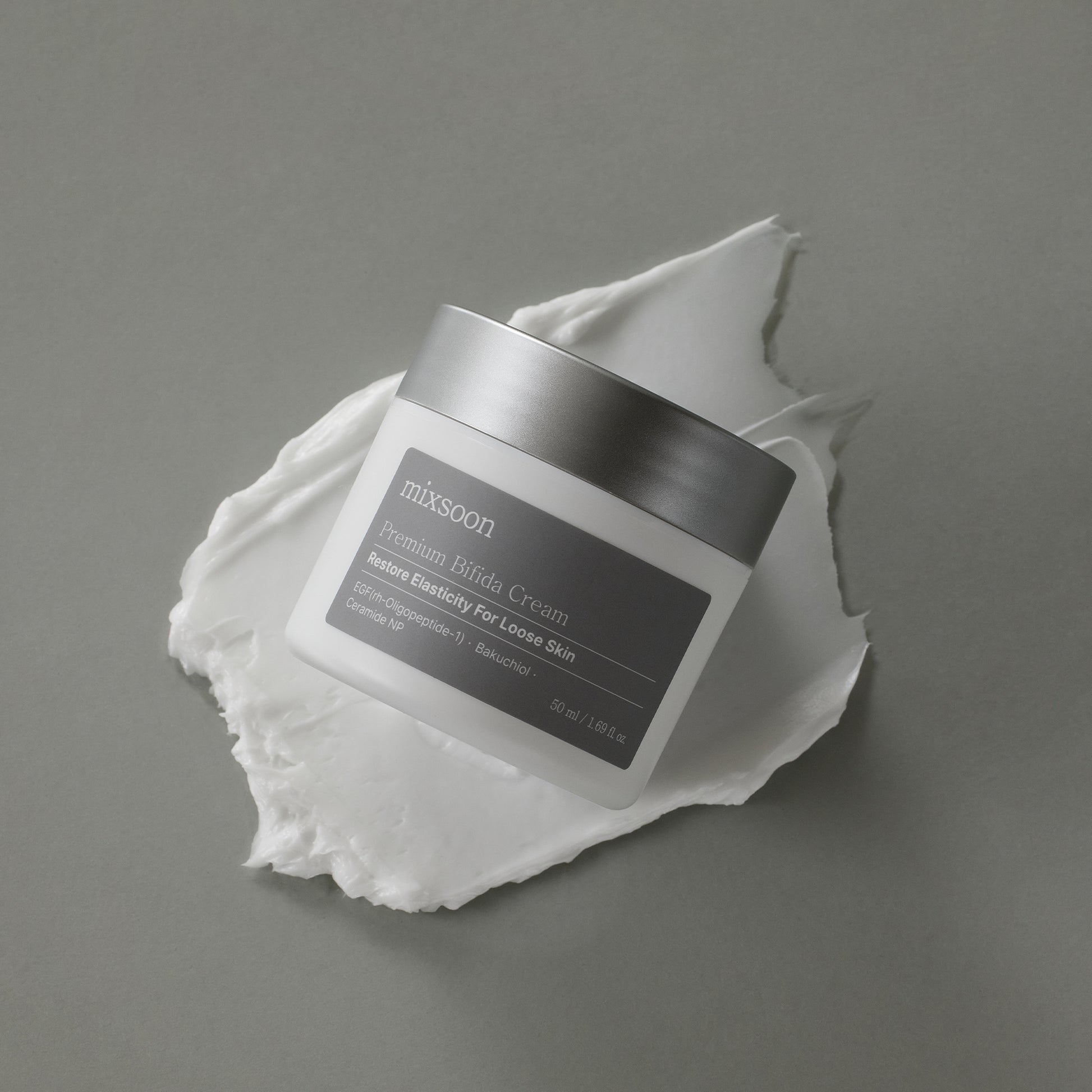 Jar of Mixsoon Premium Bifida Cream with a silver lid, placed on a smear of white cream against a gray background.