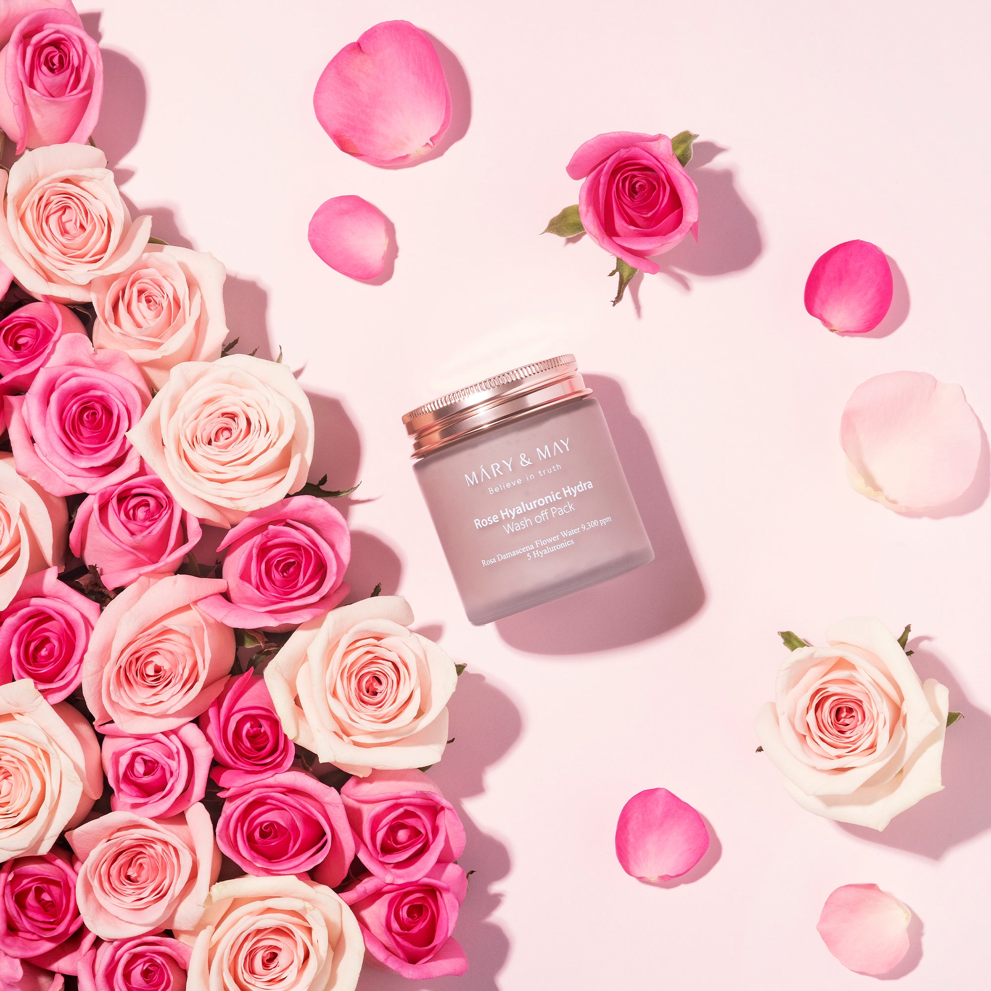 A jar of Mary & May Rose Hyaluronic Hydra Wash Off Pack placed on a light pink surface, surrounded by pink and white roses and scattered rose petals.
