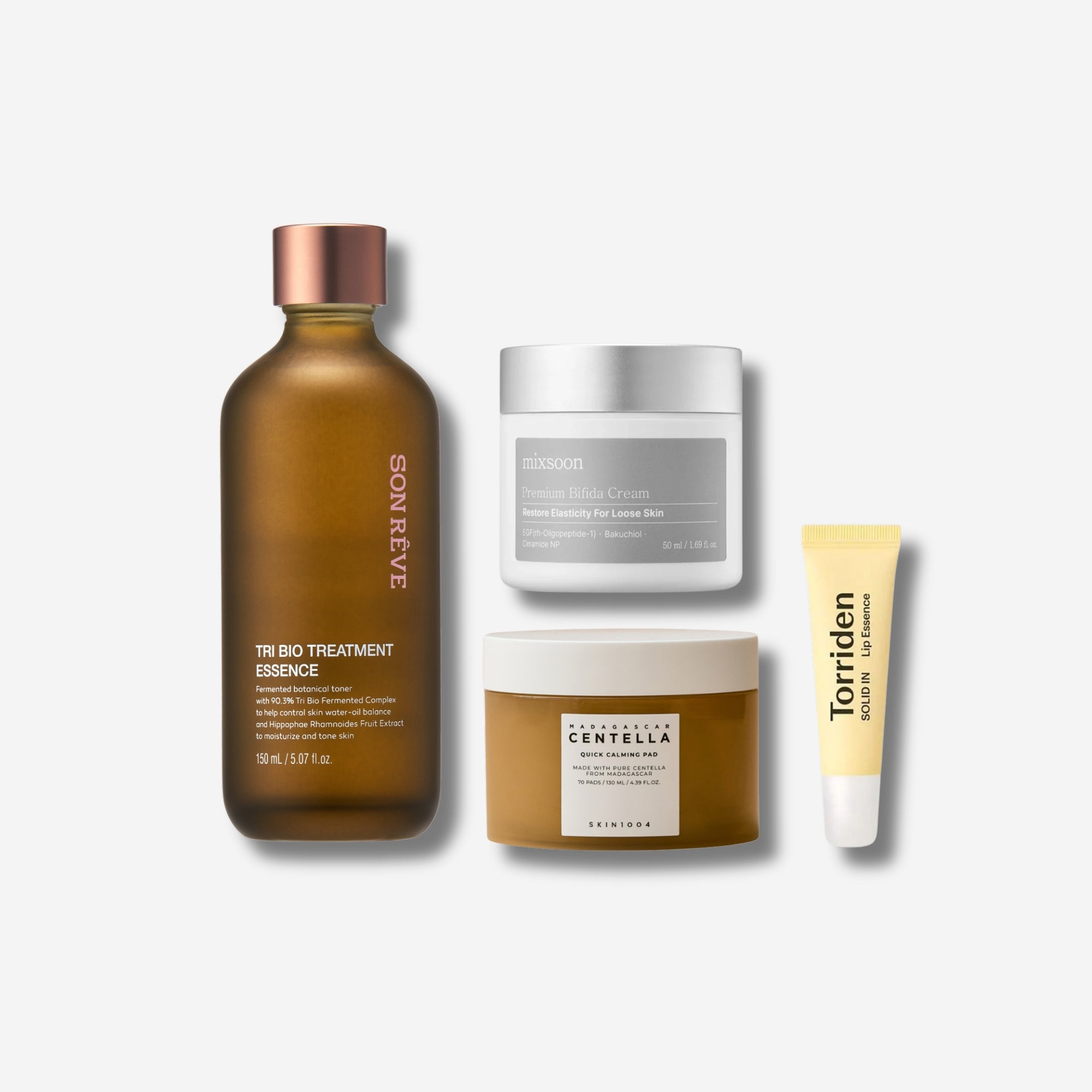 A collection of skincare products including a Son Reve Tri Bio Treatment Essence in a brown bottle with a copper cap, Mixsoon Premium Bifida Cream in a grey jar, a Skin1004 Madagascar Centella calming pad in a beige jar, and a Torriden Solid In tube in yellow.