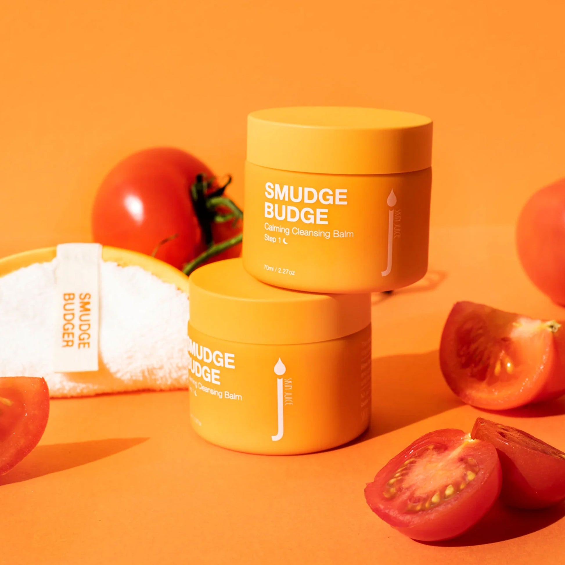 Smudge Budge Calming Cleansing Balm with cleansing pad and tomatoes on an orange background.