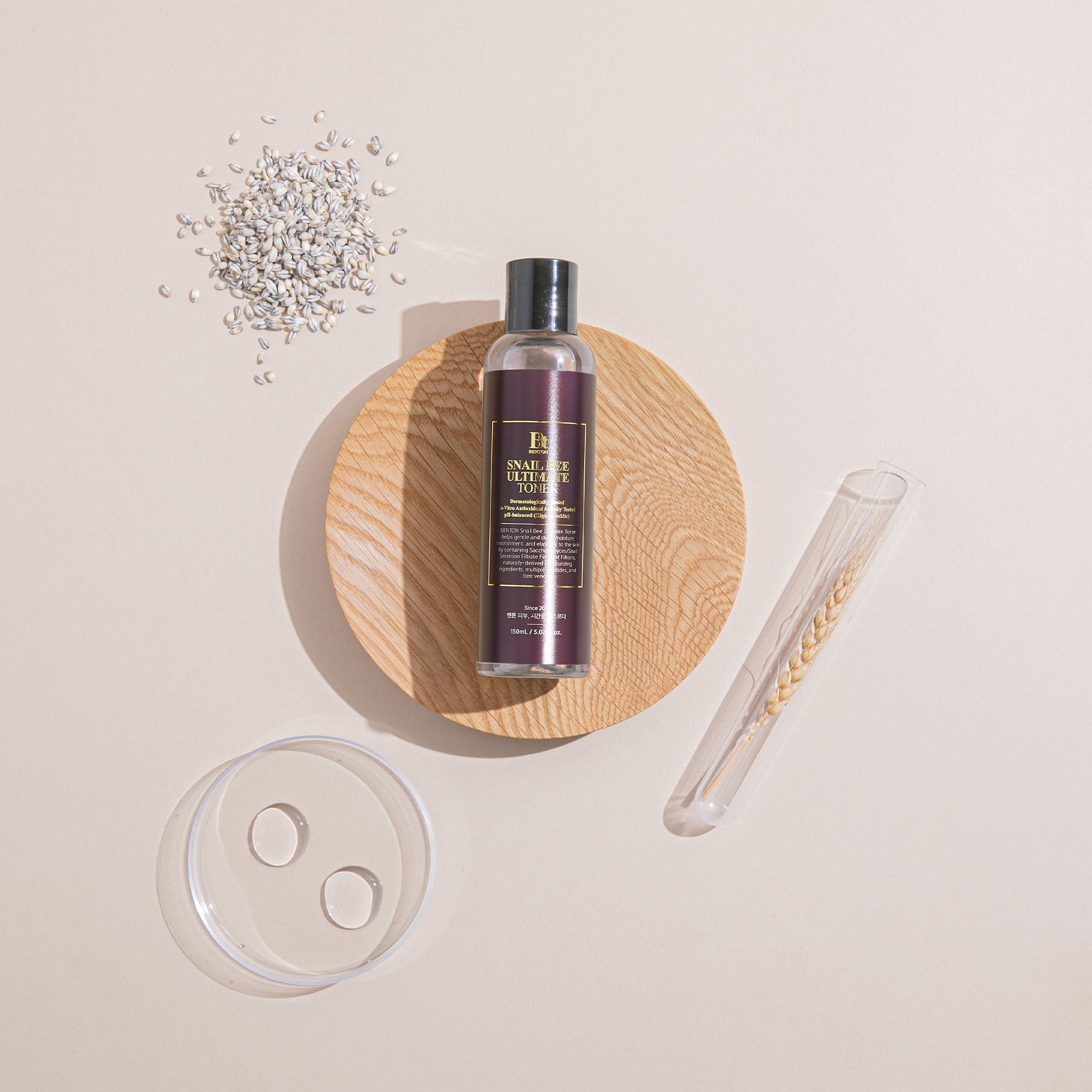 Benton Snail Bee Ultimate Toner bottle on wooden platter with grains, petri dish, and test tube background.