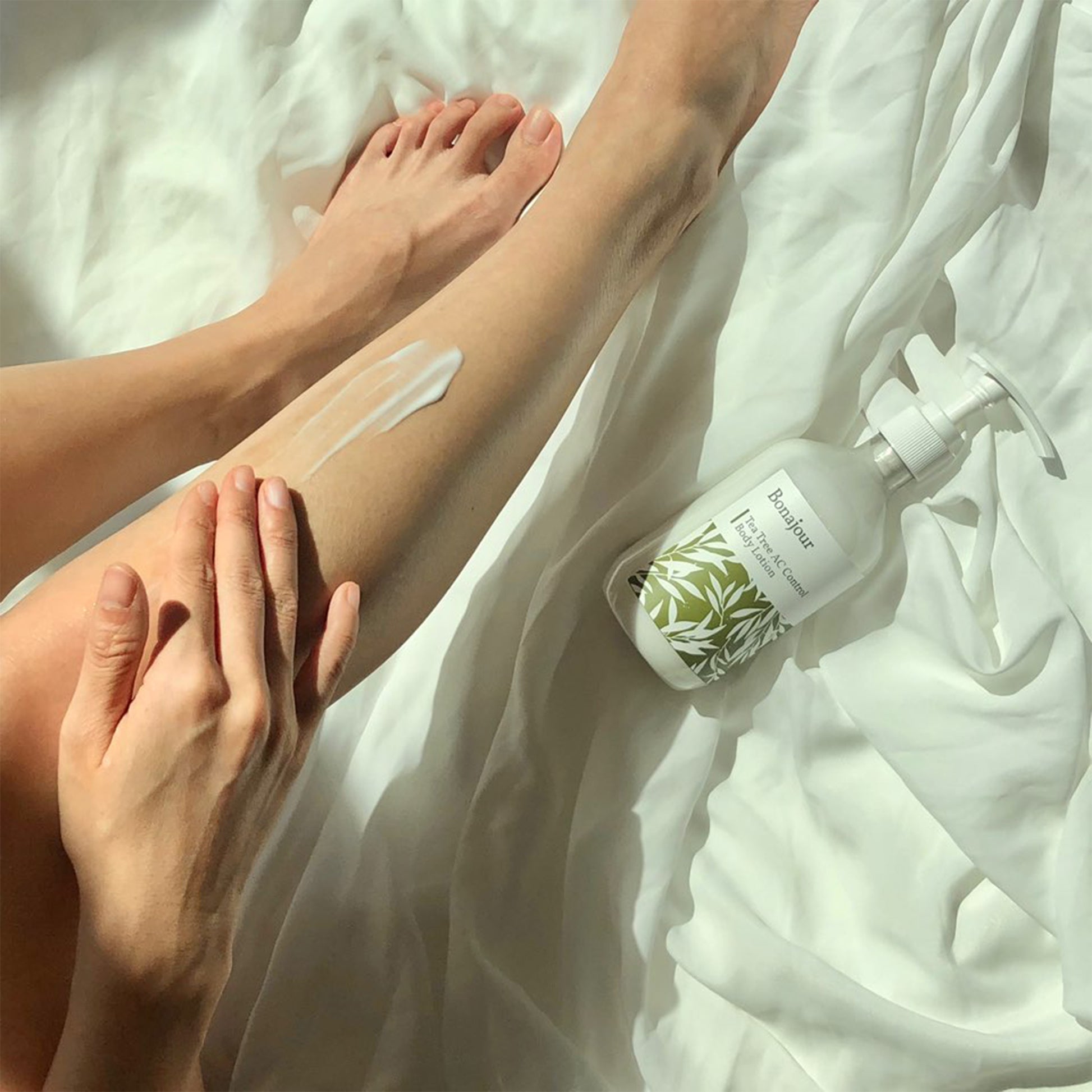 Applying Bonjour Tea Tree AC Control Body Lotion on leg, bottle nearby on white sheets