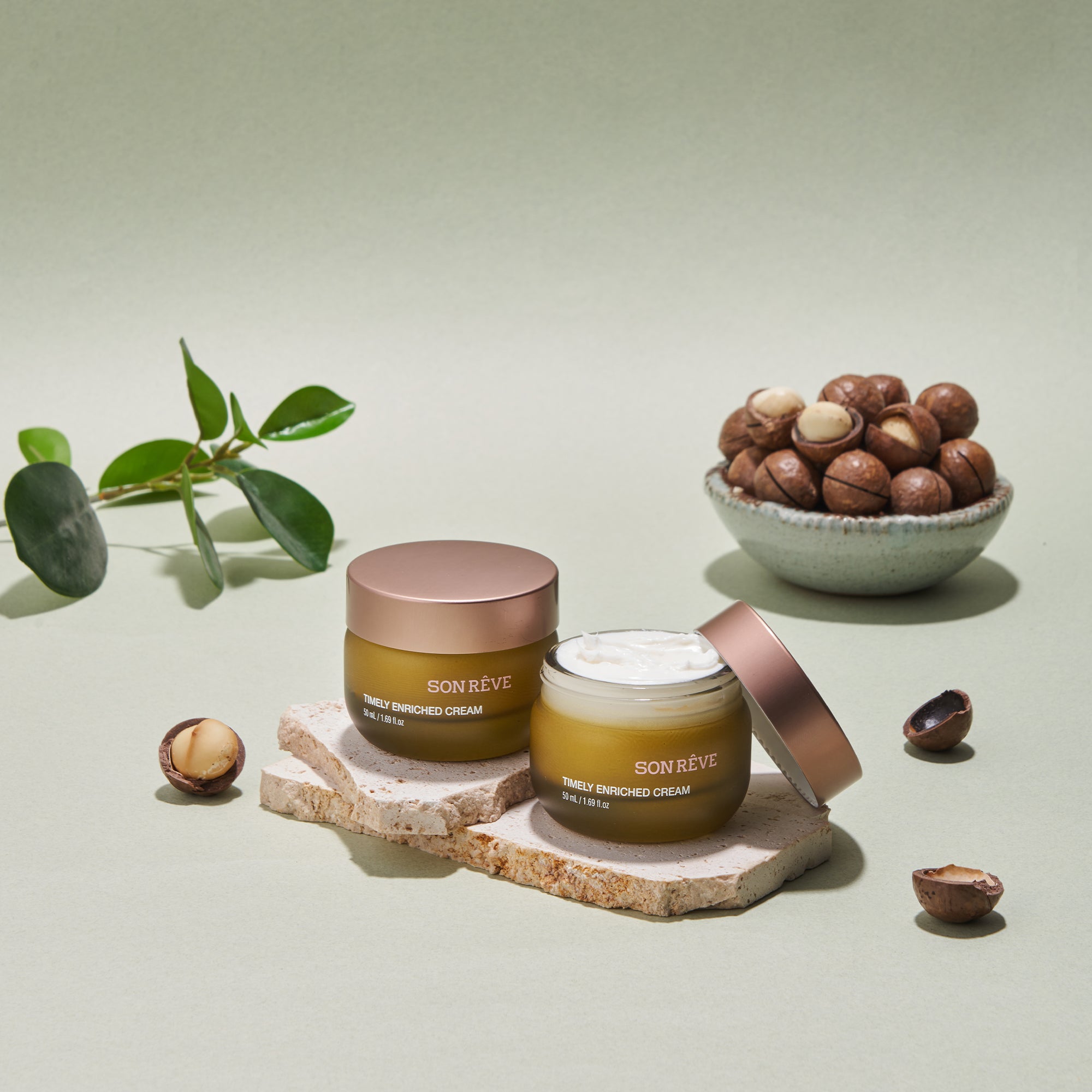 Son Réve Timely Enriched Cream jars on stone with macadamia nuts and leaves, skincare product display.