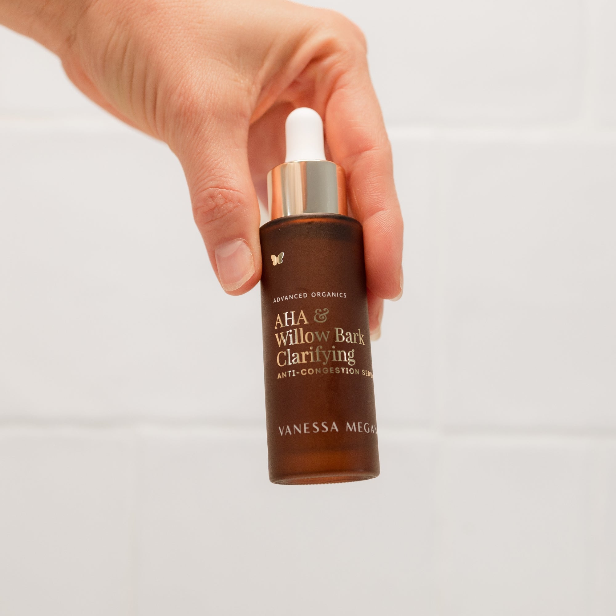 Hand holding a bottle of Vanessa Megan AHA & Willow Bark Clarifying Anti-Congestion Serum with gold cap on a light grey background