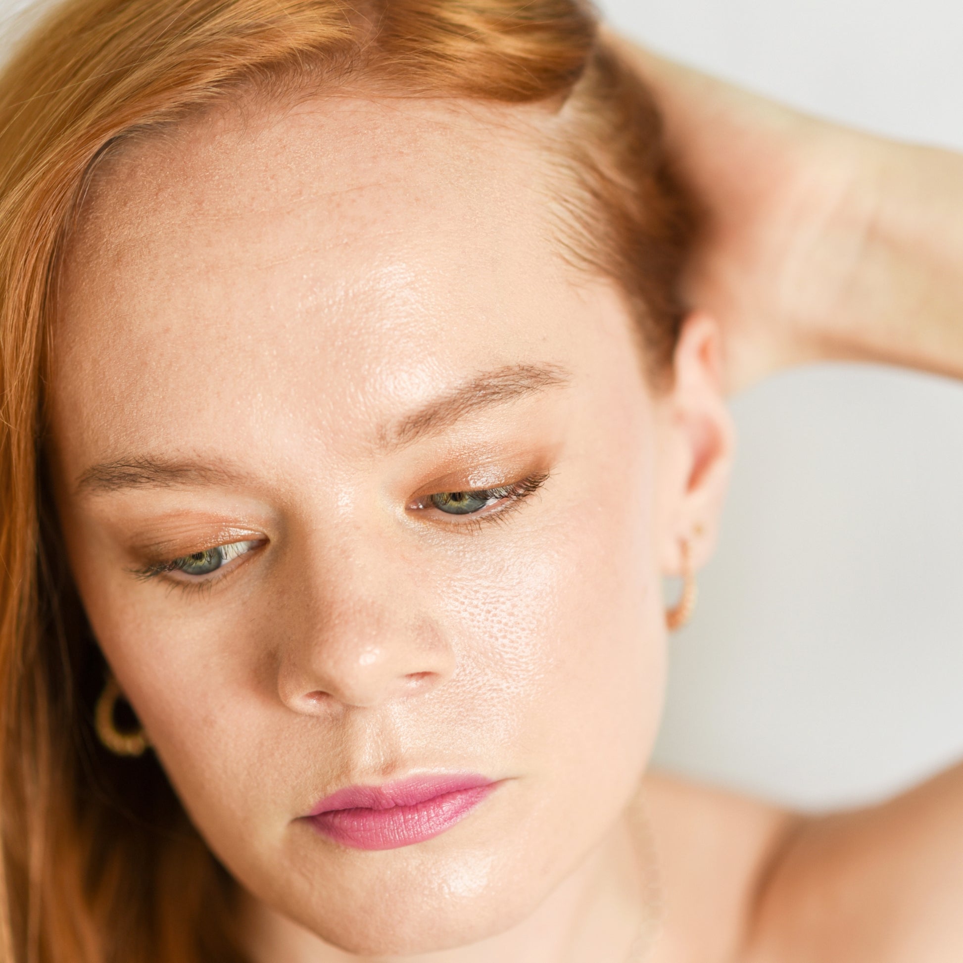 A close-up of a person wearing eye makeup using a bronzed shade, complemented with light pink lipstick. The person has their hand raised to their hair, looking downwards, showcasing a subtle and natural look.