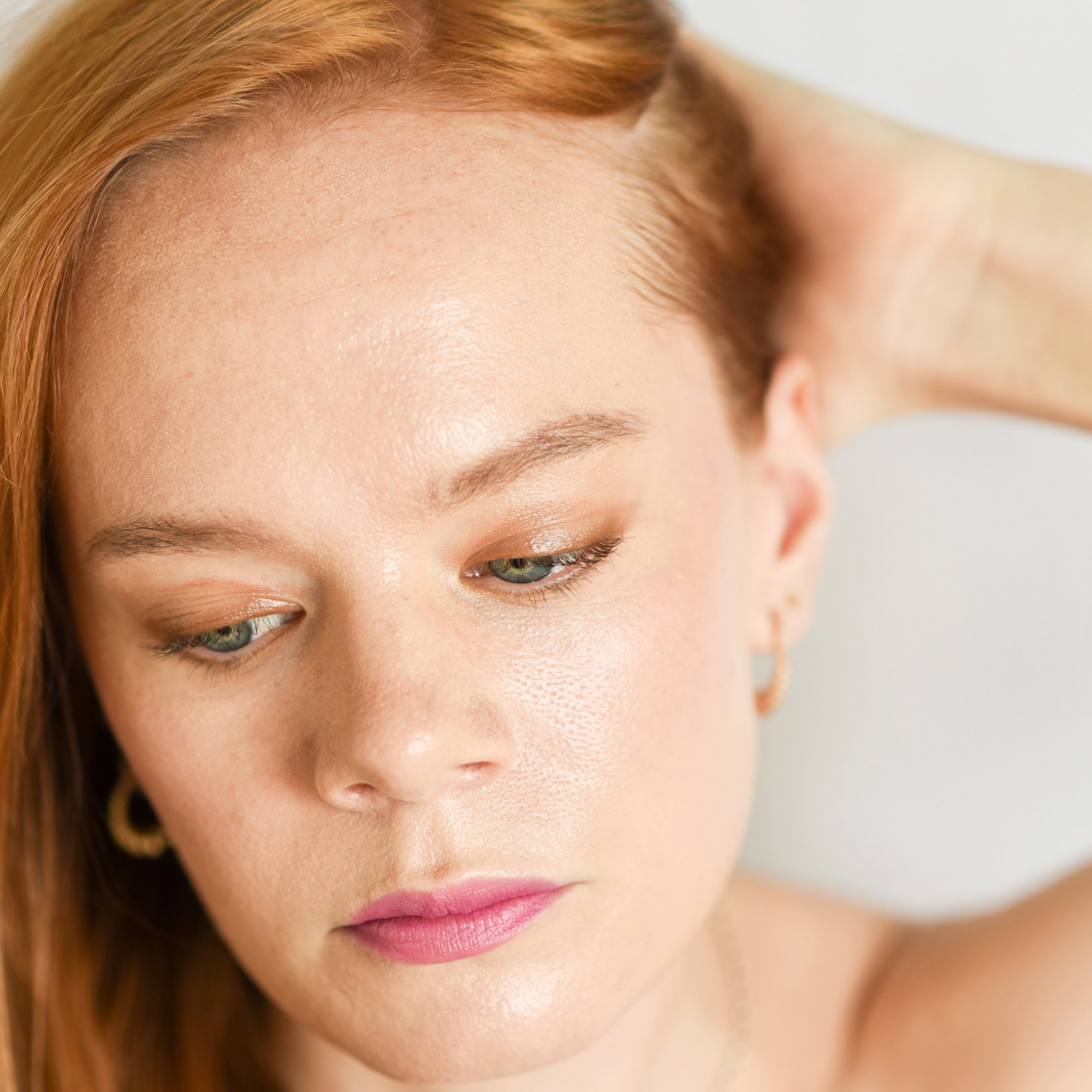 A close-up of a person wearing eye makeup using a bronzed shade, complemented with light pink lipstick. The person has their hand raised to their hair, looking downwards, showcasing a subtle and natural look.