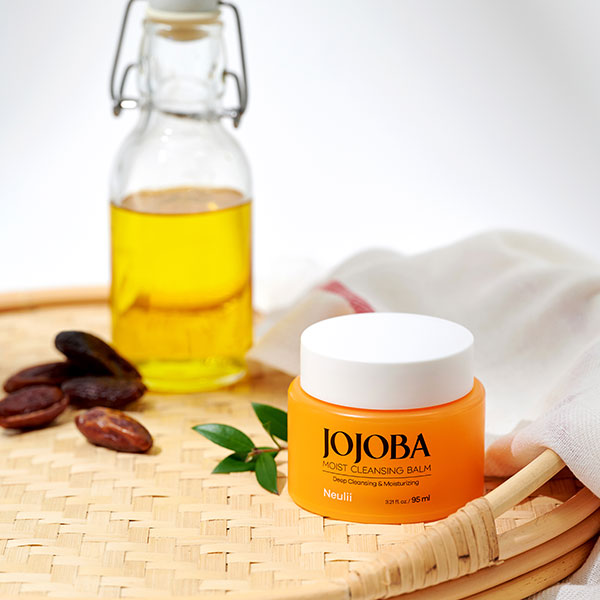 The Jojoba Moist Cleansing Balm in an orange jar, renowned as the best oil cleanser, sits beside a bottle of nourishing oil and jojoba seeds on a woven surface with a white cloth.