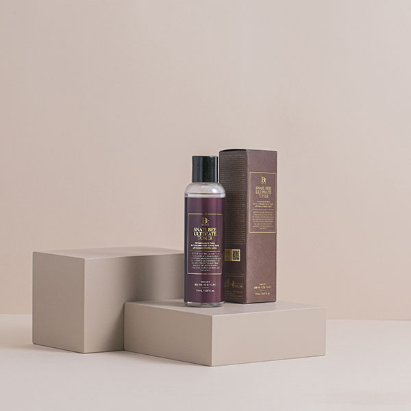 A bottle of Snail Bee Ultimate Toner and its box, both elegantly placed on beige geometric stands against a neutral background.