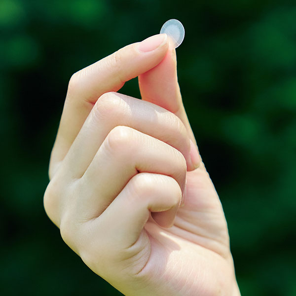 A hand holding a small, translucent Tea-Trica Spot Cover Patch between the thumb and forefinger, with a green background.