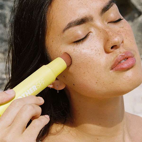 A person is using the Tinted Touch Up SPF50 sunscreen with a yellow applicator brush, enjoying the sun with closed eyes.