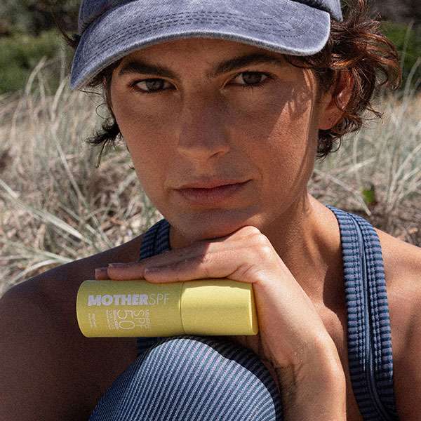 Person outdoors wearing a cap, holding a yellow bottle of Universal Face SPF50 sunscreen under their chin, looking at the camera.
